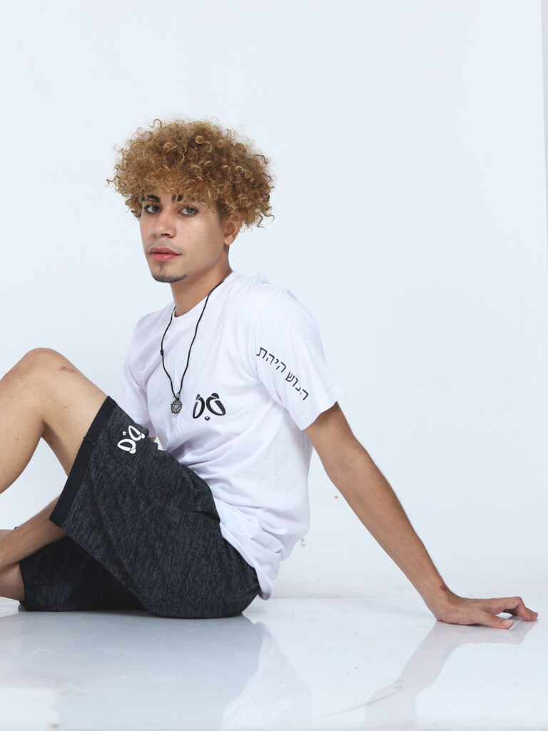 Modelo con cabello rizado rubio y pecas, usando camiseta blanca Aypanda con escritura en manga y shorts grises, sentado para foto de moda juvenil y auténtica.