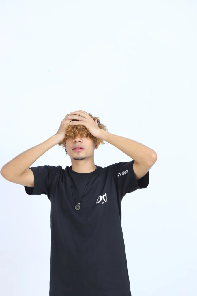 Joven modelo con camiseta negra Aypanda, cabello rizado rubio y manos en la cabeza, expresando un estilo relajado y libre característico de la marca.