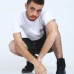 Modelo joven con cabello rizado y pecas, en cuclillas, vistiendo camiseta blanca Aypanda con logo y shorts verde oliva, transmitiendo una pose auténtica y dinámica.
