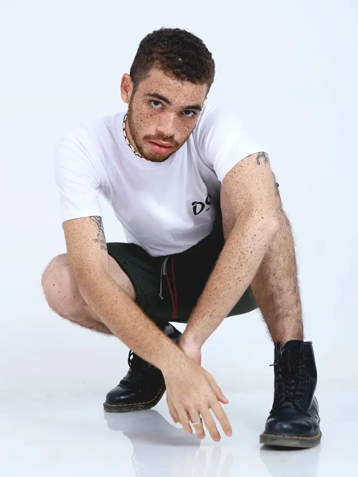 Modelo joven con cabello rizado y pecas, en cuclillas, vistiendo camiseta blanca Aypanda con logo y shorts verde oliva, transmitiendo una pose auténtica y dinámica.