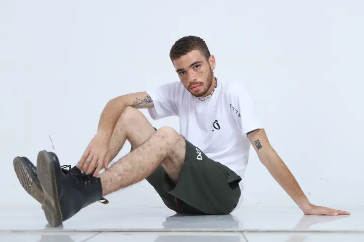 Joven modelo con camiseta blanca Aypanda y shorts verde oliva con cordón rojo, posando sentado para un look casual y auténtico de la marca.