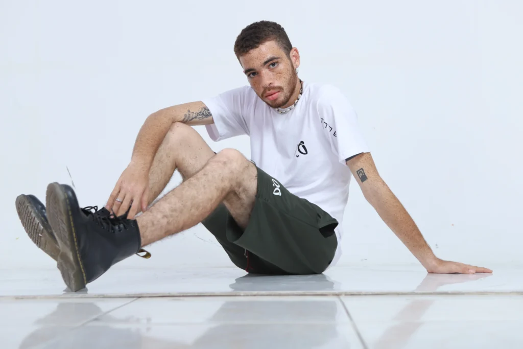 Joven modelo con camiseta blanca Aypanda y shorts verde oliva con cordón rojo, posando sentado para un look casual y auténtico de la marca.