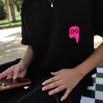 Persona usando camiseta negra Fruit of the Forest de Aypanda con logo rosa en el pecho, sentada al aire libre.
